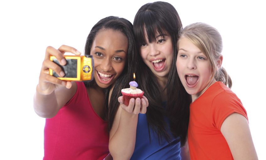 Birthday celebration photograph with candle on cakes for three beautiful young teenager girl friends a blonde caucasian, an oriental Japanese and an African American girl holding the camera.